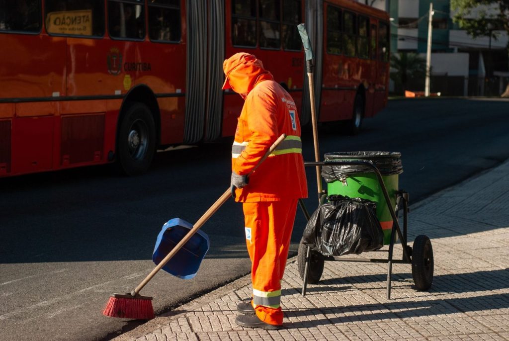 Profissional uniformizado varre uma calçada próximo a um ônibus em uma área urbana.